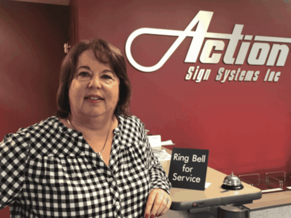 Portrait of a smiling woman standing at the reception desk of Action Sign Systems Inc, with the company logo on a red wall behind her and a small “Ring Bell for Service” sign on the counter.
