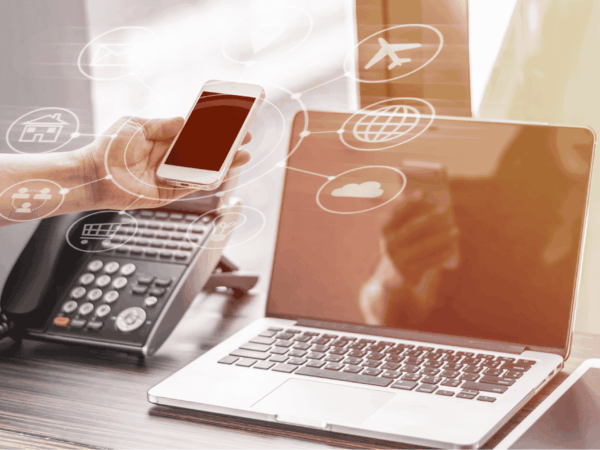 Person holding a smartphone near a laptop and office desk phone, with digital icons representing cloud computing, internet, travel, email, and shopping overlaid, symbolizing modern business communication technology.
