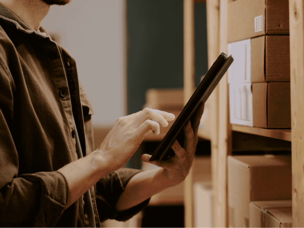 Employee using a tablet to track IT hardware inventory on storage shelves as part of lifecycle management and asset tracking.