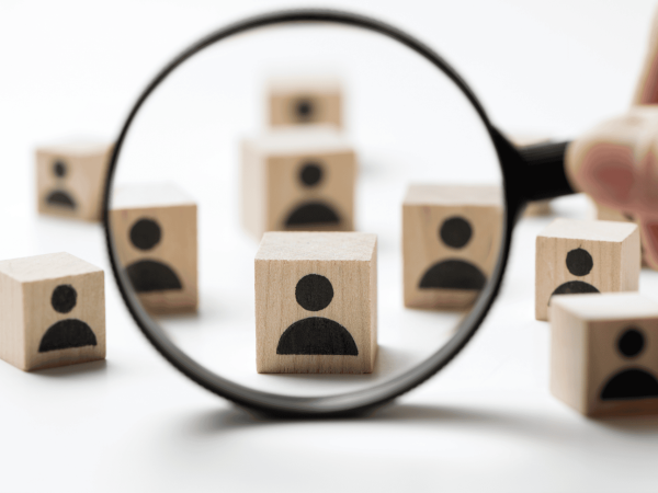 Magnifying glass focusing on a wooden block with a person icon – identifying user accounts for immediate deactivation during off-boarding.