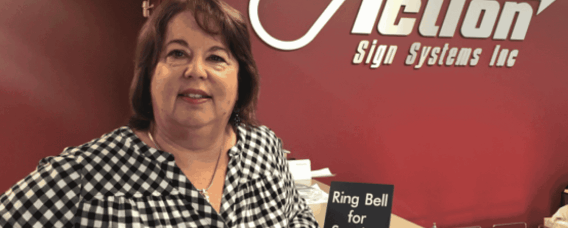 Portrait of a smiling woman standing at the reception desk of Action Sign Systems Inc, with the company logo on a red wall behind her and a small “Ring Bell for Service” sign on the counter.