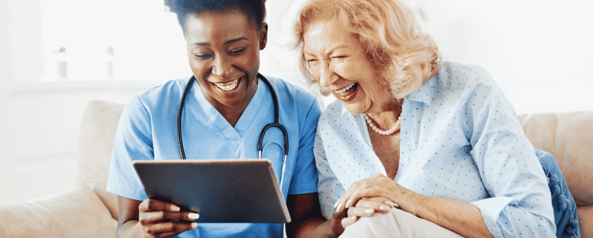 Home health caregiver reviewing a tablet with a laughing senior client on a couch, representing EMR and EVV technology used in home-based care