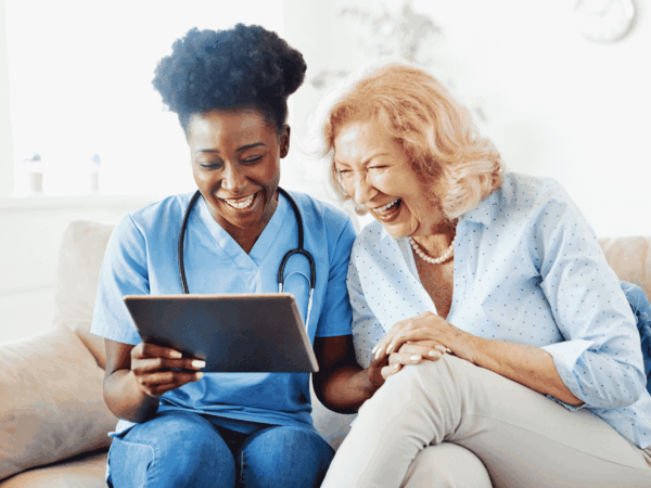 Home health caregiver reviewing a tablet with a laughing senior client on a couch, representing EMR and EVV technology used in home-based care