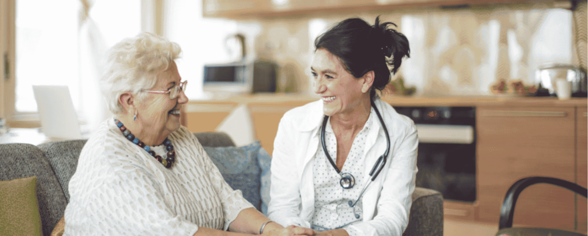 Older woman smiling and holding hands with a healthcare professional during an in-home visit.