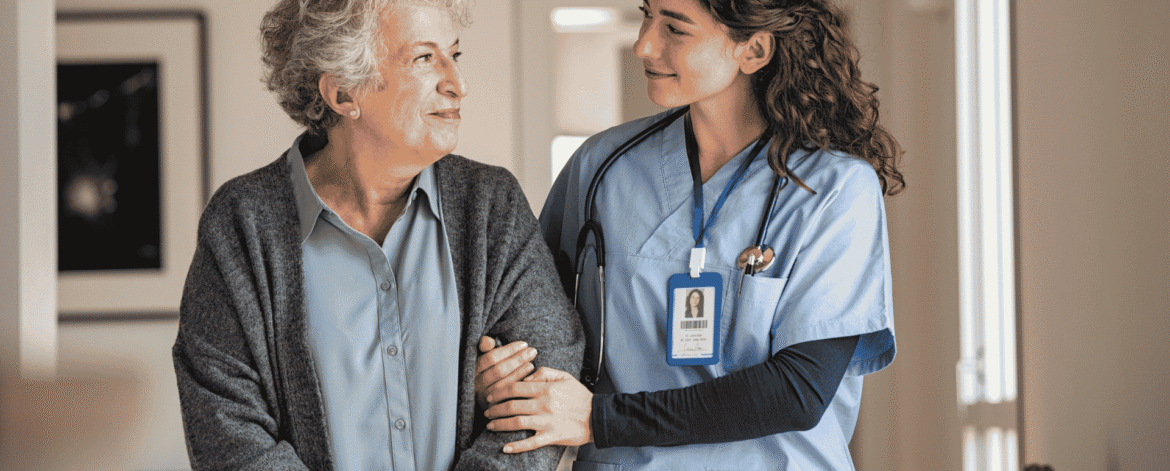Image 1_10.19.26 a nurse in blue scrubs with stethoscope and ID badge holding the arm of an older woman in a gray cardigan, standing in her home