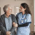 a nurse in blue scrubs with stethoscope and ID badge holding the arm of an older woman in a gray cardigan, standing in her home