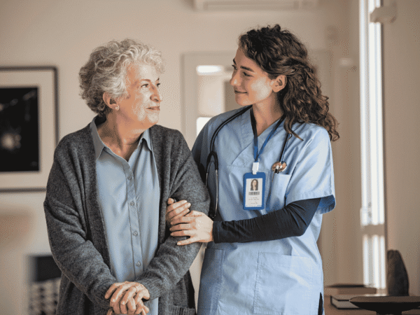 a nurse in blue scrubs with stethoscope and ID badge holding the arm of an older woman in a gray cardigan, standing in her home