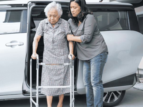 Caregiver steadying an older woman with a walker beside a van after a home visit, the care that precedes questions about HIPAA rules for deceased hospice patients.