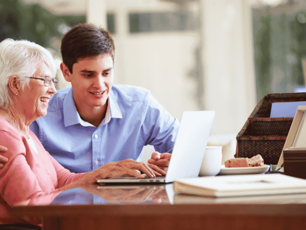 Older adult and a younger man reviewing a laptop together at her kitchen table, the setting behind decisions about outsourcing HIPAA compliance for home health agencies.