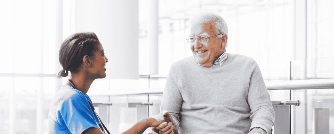 Nurse in blue scrubs holding hands with an older man in a wheelchair beside tall windows, the client relationships that cybersecurity for home care agencies exists to protect.