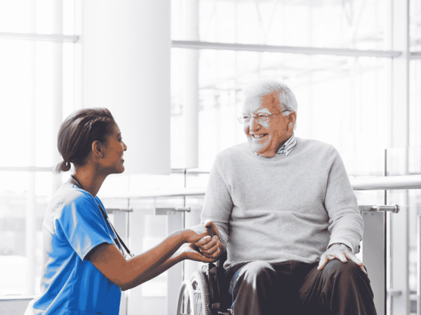 Nurse in blue scrubs holding hands with an older man in a wheelchair beside tall windows, the client relationships that cybersecurity for home care agencies exists to protect.