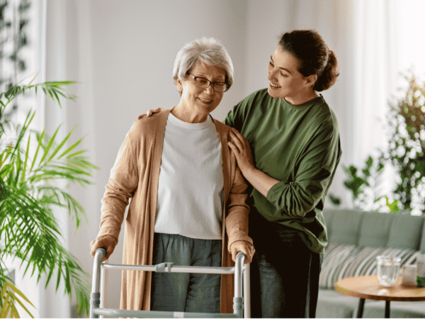 Image 2_8.26.26 Older woman using a walker, steadied by an aide in a sunlit living room where the field devices in an ePHI asset inventory for home health agencies are actually used.