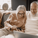 older couple on a couch looking at a smartphone together, laptop open in front of them