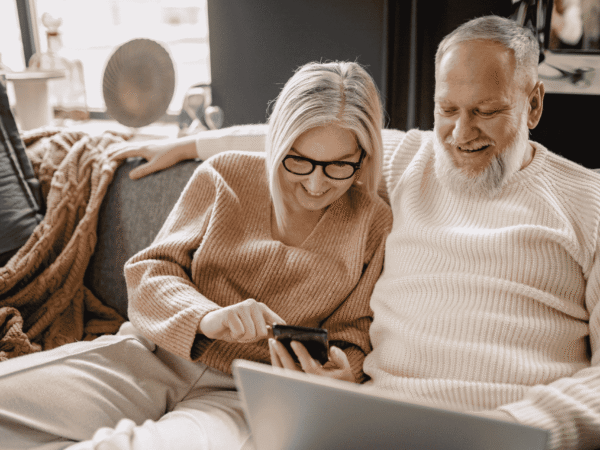older couple on a couch looking at a smartphone together, laptop open in front of them