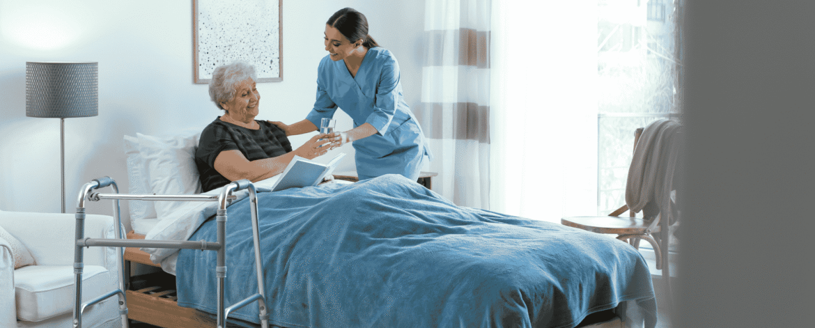 a caregiver in blue scrubs handing a glass of water to an older woman reading in bed, with a walker in the foreground