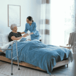 a caregiver in blue scrubs handing a glass of water to an older woman reading in bed, with a walker in the foreground