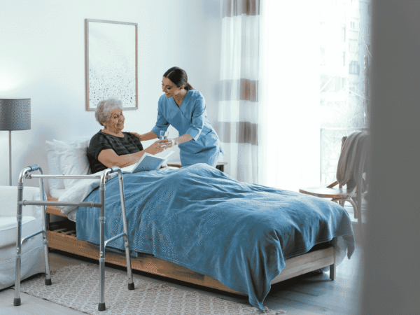 a caregiver in blue scrubs handing a glass of water to an older woman reading in bed, with a walker in the foreground