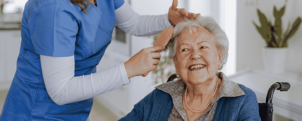 Home health aide in blue scrubs brushing a seated client's hair during a home visit, the care setting behind how to report a medical information breach to CDPH.