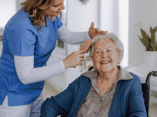 Image 2_9.16.26 Home health aide in blue scrubs brushing a seated client's hair during a home visit, the care setting behind how to report a medical information breach to CDPH.