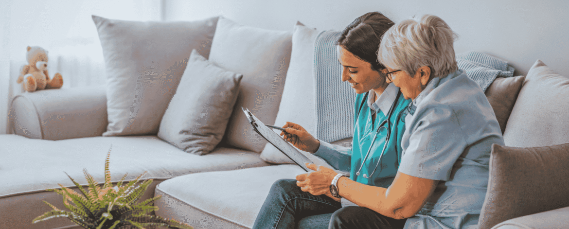 Clinician in scrubs reviewing patient notes beside an older adult on a couch, the documentation routine behind how often home health agencies need a HIPAA risk assessment.