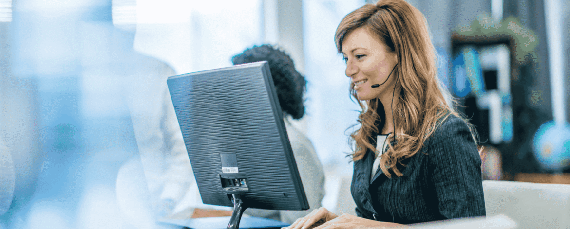 Support agent in a headset typing at a desktop computer in a bright office, fielding questions such as whether public Wi-Fi is a HIPAA violation for home health clinicians.
