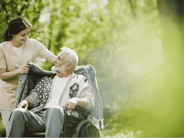 Image 3 Caregiver wrapping a blanket around an older man in a wheelchair outdoors during a hospice visit, where ePHI travels on field devices subject to Security Rule safeguards.