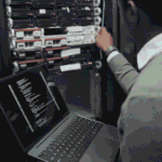 technician holding an open laptop while adjusting a server in a dark equipment rack