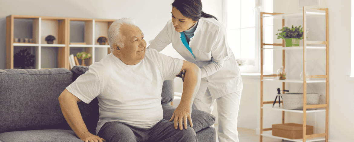 Caregiver in a white uniform helping an older man rise from a sofa in a sunlit living room, the non-medical personal care that decides whether HIPAA applies to home care agencies.
