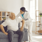 Caregiver in a white uniform helping an older man rise from a sofa in a sunlit living room, the non-medical personal care that decides whether HIPAA applies to home care agencies.