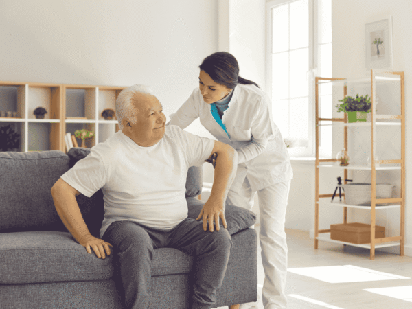 Caregiver in a white uniform helping an older man rise from a sofa in a sunlit living room, the non-medical personal care that decides whether HIPAA applies to home care agencies.