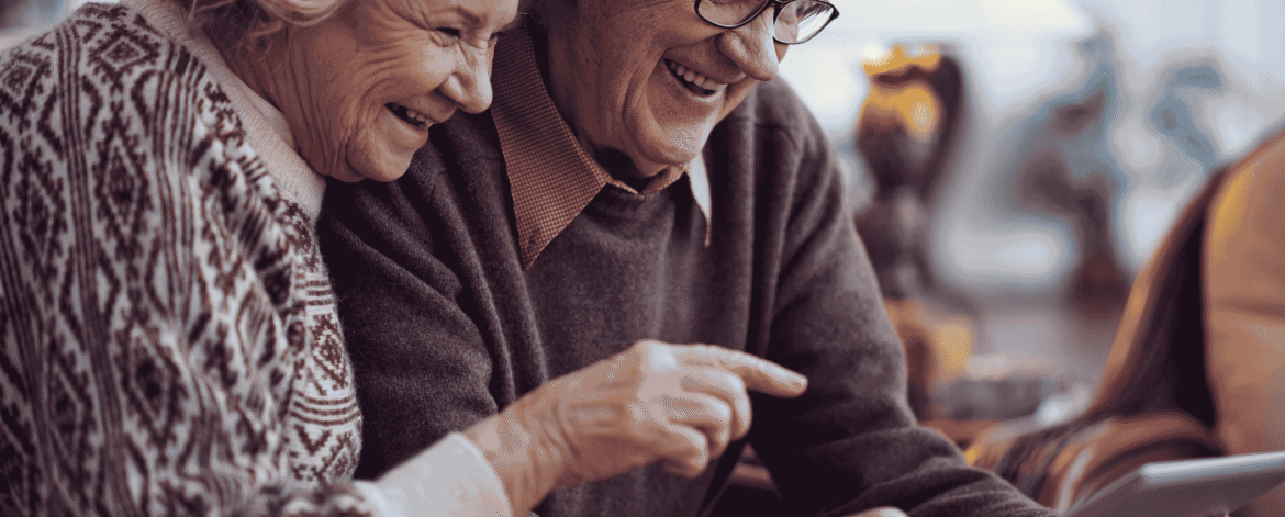 Image 3_10.23.26 an older couple on a couch laughing as the man points at a tablet