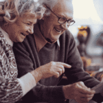 an older couple on a couch laughing as the man points at a tablet