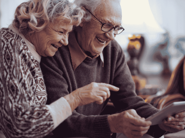 an older couple on a couch laughing as the man points at a tablet